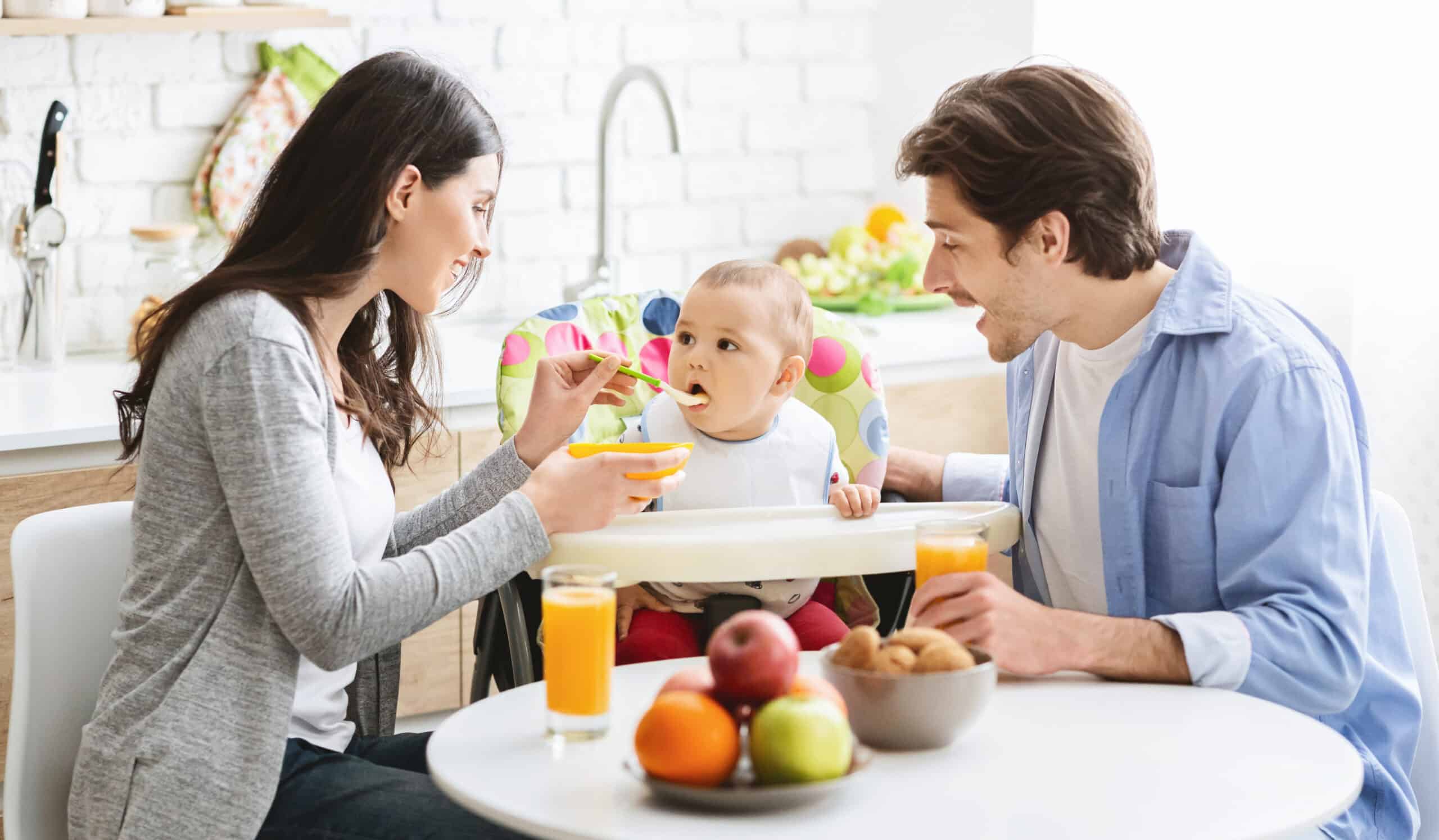 Parents feeding baby in high chair with spoon and bowl, breakfast table with fruits and juice in foreground.