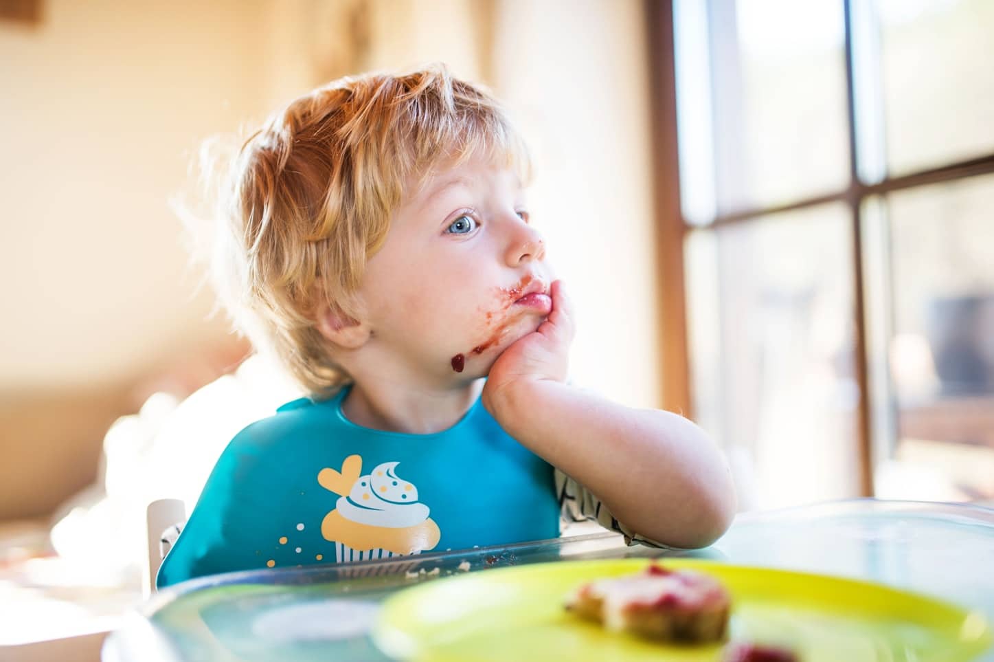 Toddler with food-stained face gazes out window, wearing a cupcake-themed bib.