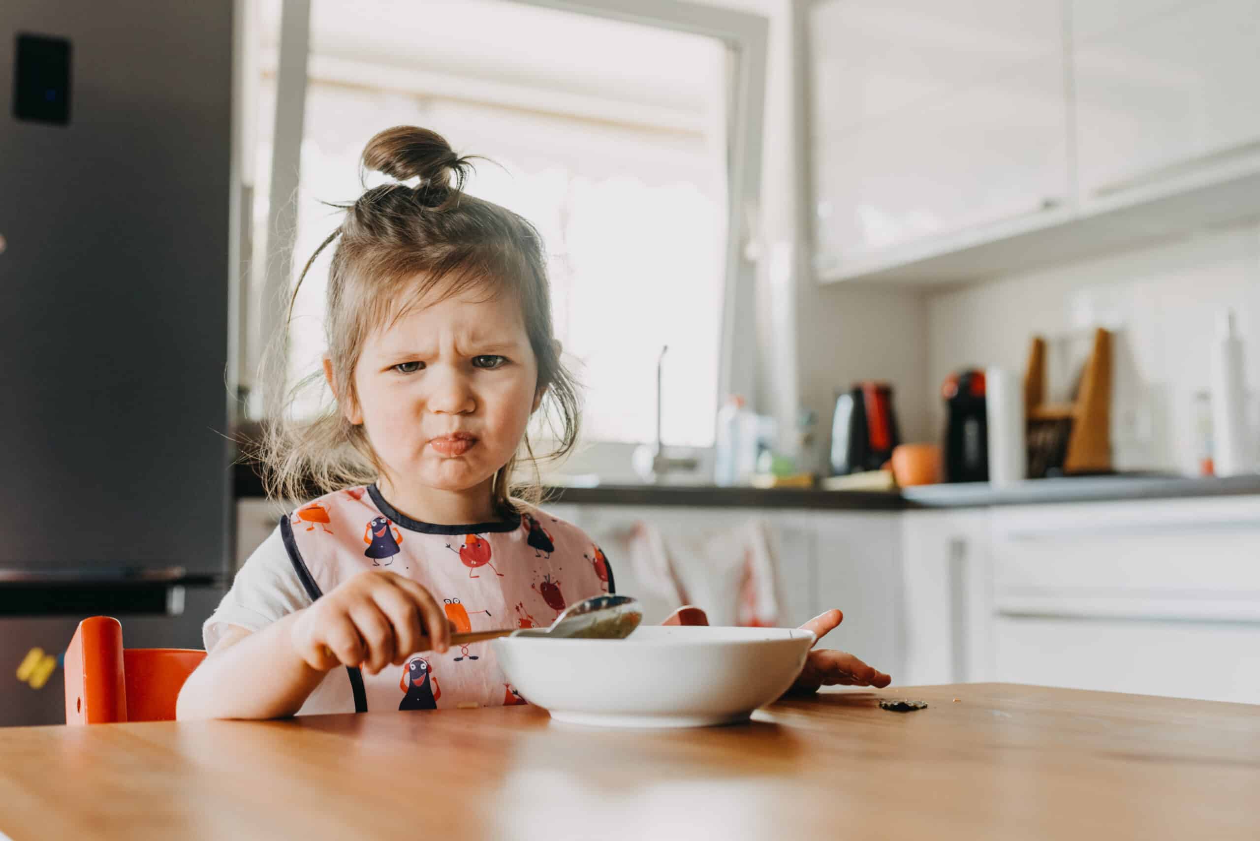Toddler in a bib frowns at a bowl of food in a kitchen.