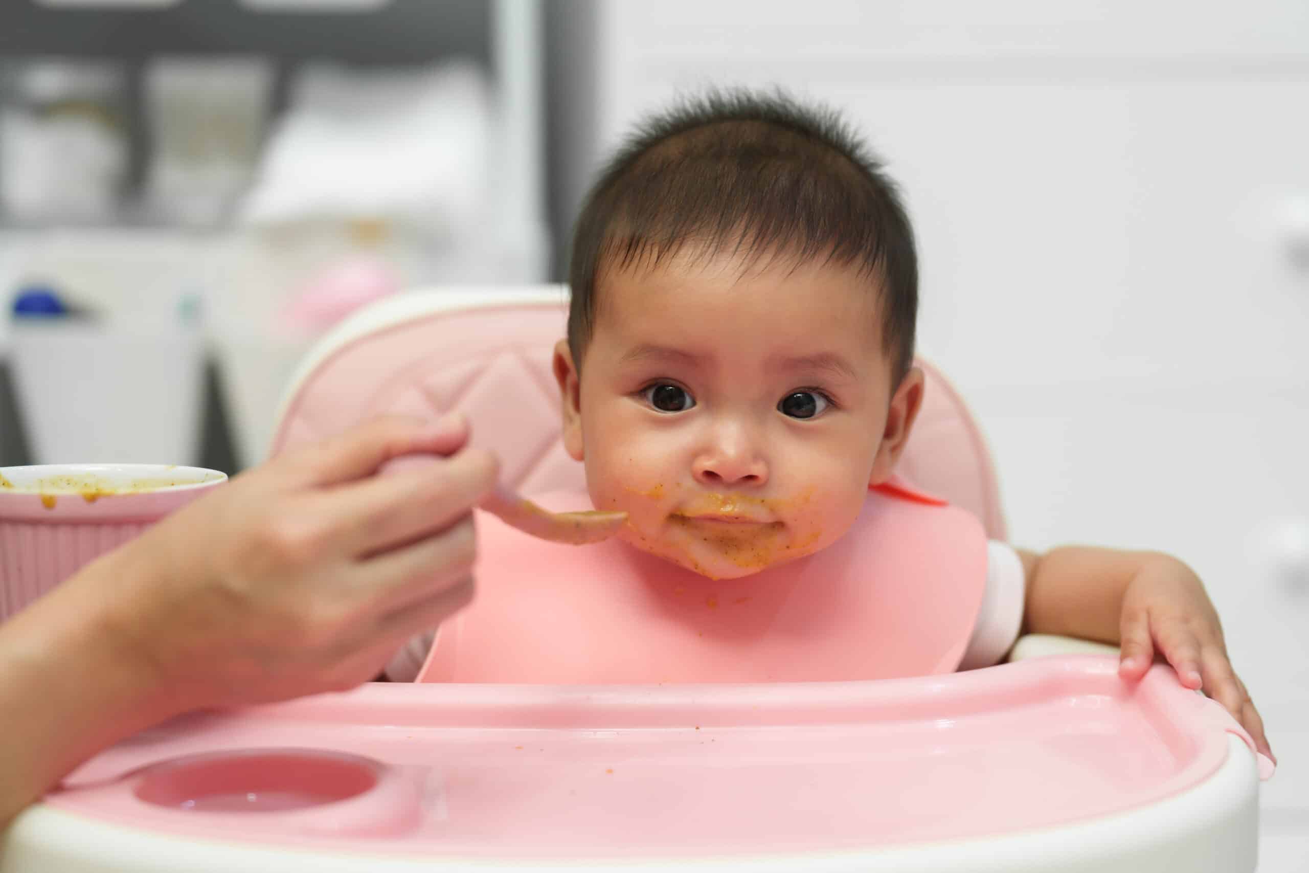 Baby in highchair eating puree, with a spoonful approaching, wearing a pink bib.
