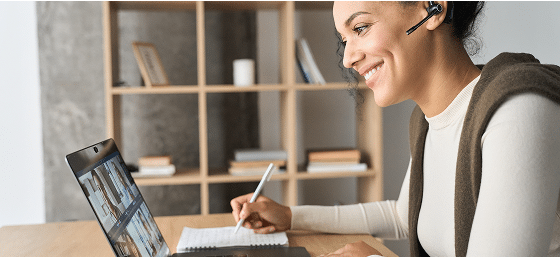 Smiling person with headset participating in a virtual meeting on a laptop, taking notes.