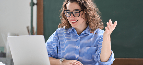 Smiling woman in glasses gestures while looking at a laptop, sitting in front of a chalkboard.