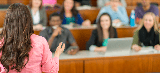 A woman in a pink shirt speaks to a classroom of attentive students.